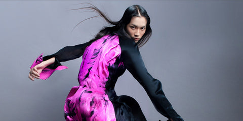 A woman with long dark hair poses in a dynamic crouch, wearing a striking black dress with vivid pink and purple patterns against a plain gray background.