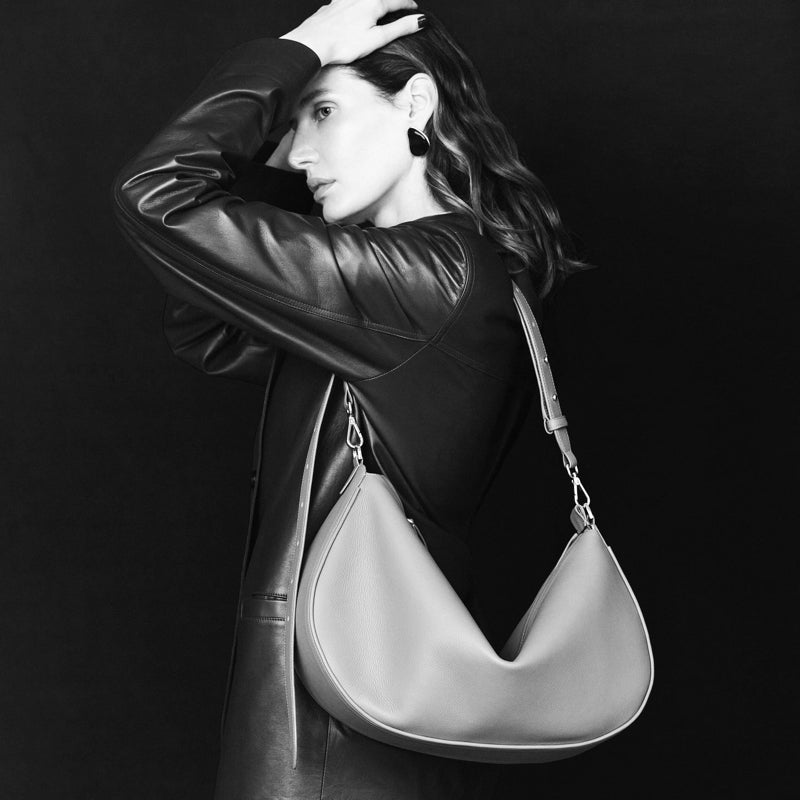 Black and white photo of a woman in a leather jacket posing with the large crescent-shaped Denise Bag | Gray by VIKELE STUDIO. The dark background accentuates her striking outfit and this luxury handbag.