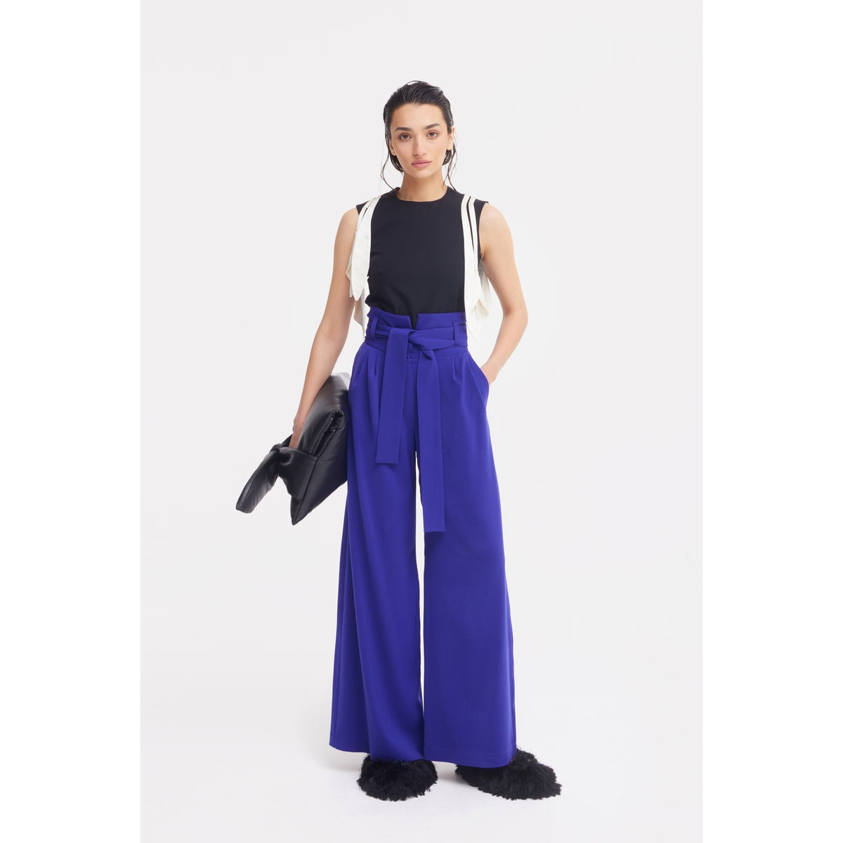 A woman stands against a white background in a black sleeveless top, JULIA ALLERT Blue High Waist Long Wide Leg Trousers with a tie belt, furry black shoes, a white sweater over her shoulders, and holding a large black clutch.