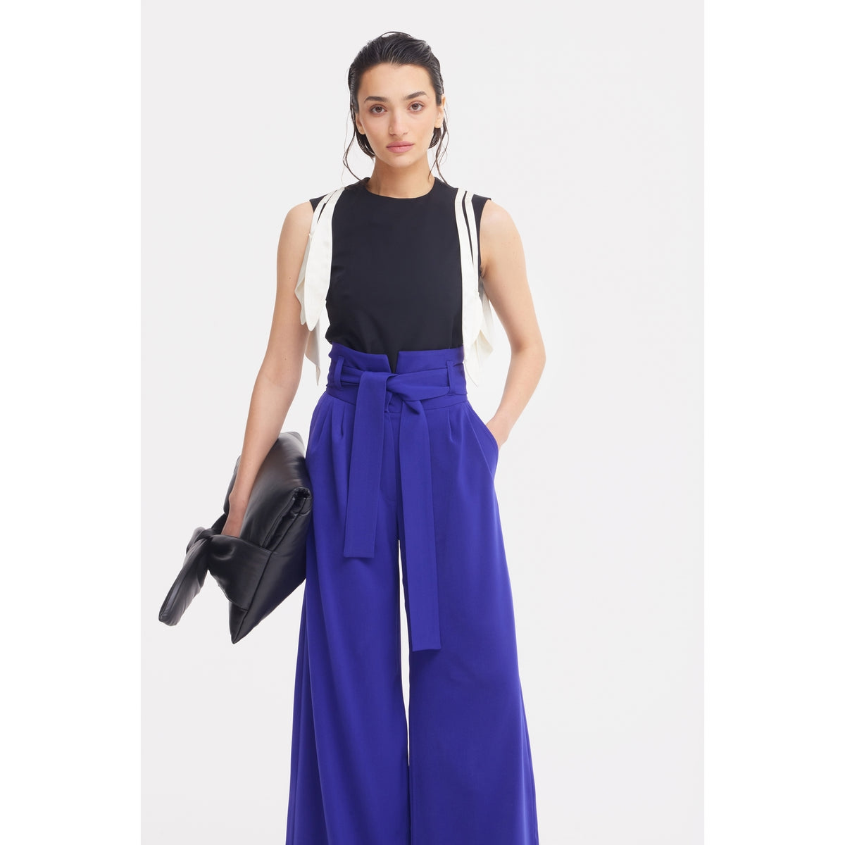 A woman faces forward in JULIA ALLERT Blue High Waist Long Wide Leg Trousers, a sleeveless black top, and a white garment over her shoulders, holding a large black clutch bag.