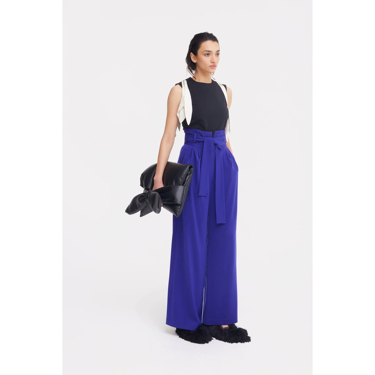 A woman stands against a white background wearing a sleeveless black top, JULIA ALLERT Blue High Waist Long Wide Leg Trousers with a belt, holding a black clutch with a large bow, and black fuzzy shoes.