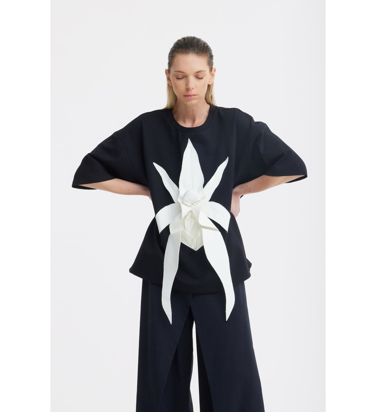 A woman with light brown hair wears the JULIA ALLERT Relaxed Fit Knit Top in black, featuring a large white sculptural floral appliqué on the front, paired with wide-leg black pants, standing against a plain white background.