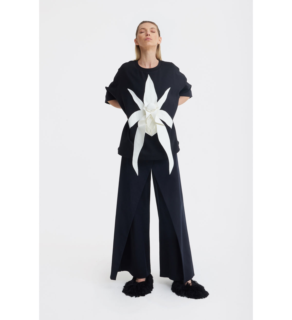 A woman stands with her hands behind her back, wearing the JULIA ALLERT Relaxed Fit Knit Top in black paired with wide-legged black pants and black fuzzy slippers, set against a plain white background.