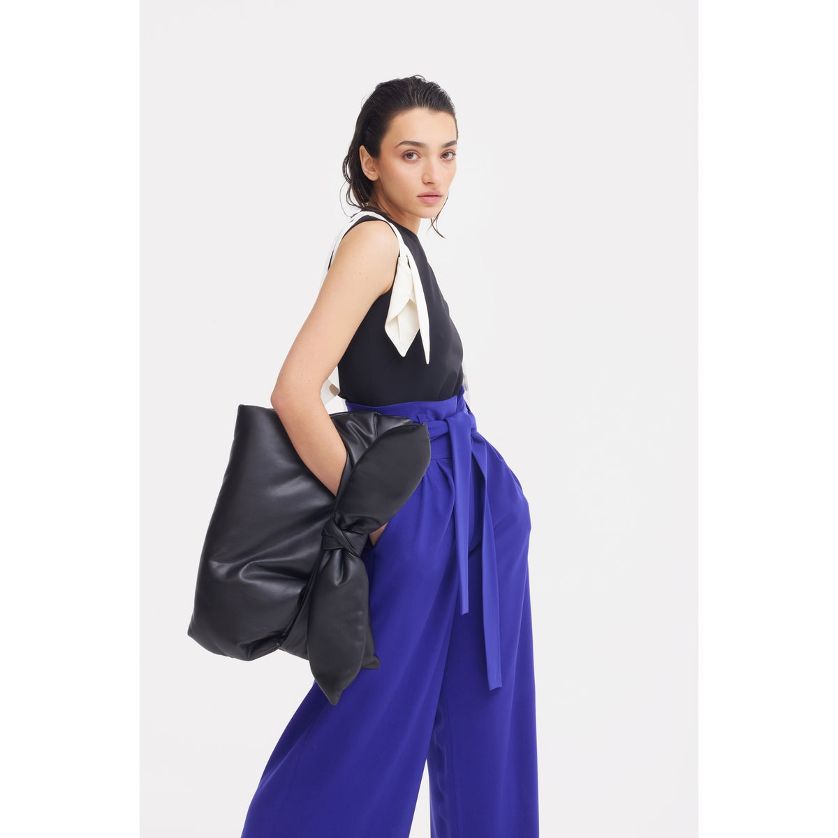 Woman poses against a white background wearing sleeveless black top with white bow, JULIA ALLERT Blue High Waist Long Wide Leg Trousers with sash, and holding large black clutch bag with a bow.