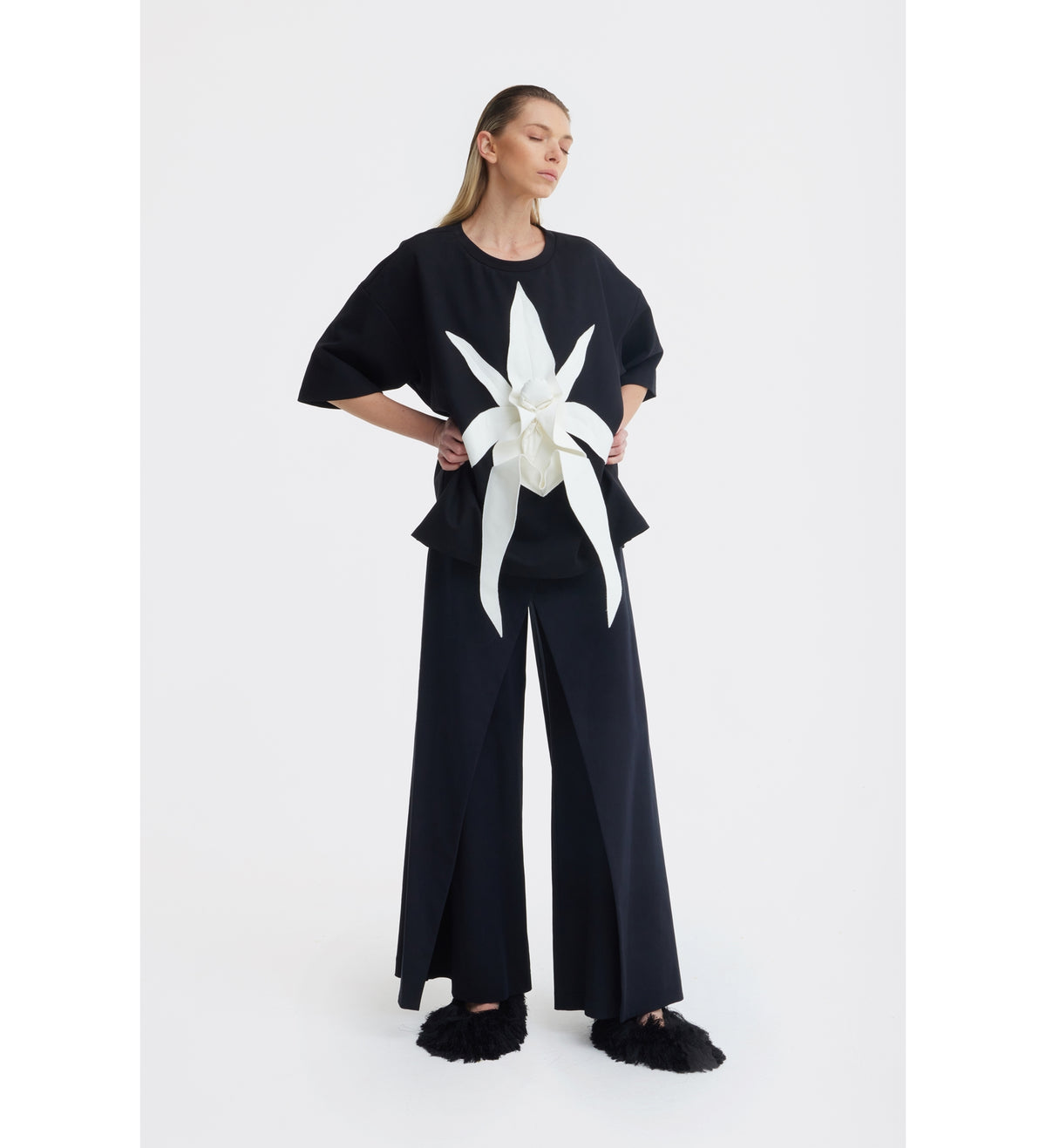 A woman stands against a plain white background in the JULIA ALLERT Relaxed Fit Knit Top - Black with wide-leg pants, showcasing a bold sculptural floral appliqué on the front and paired with black furry slippers.