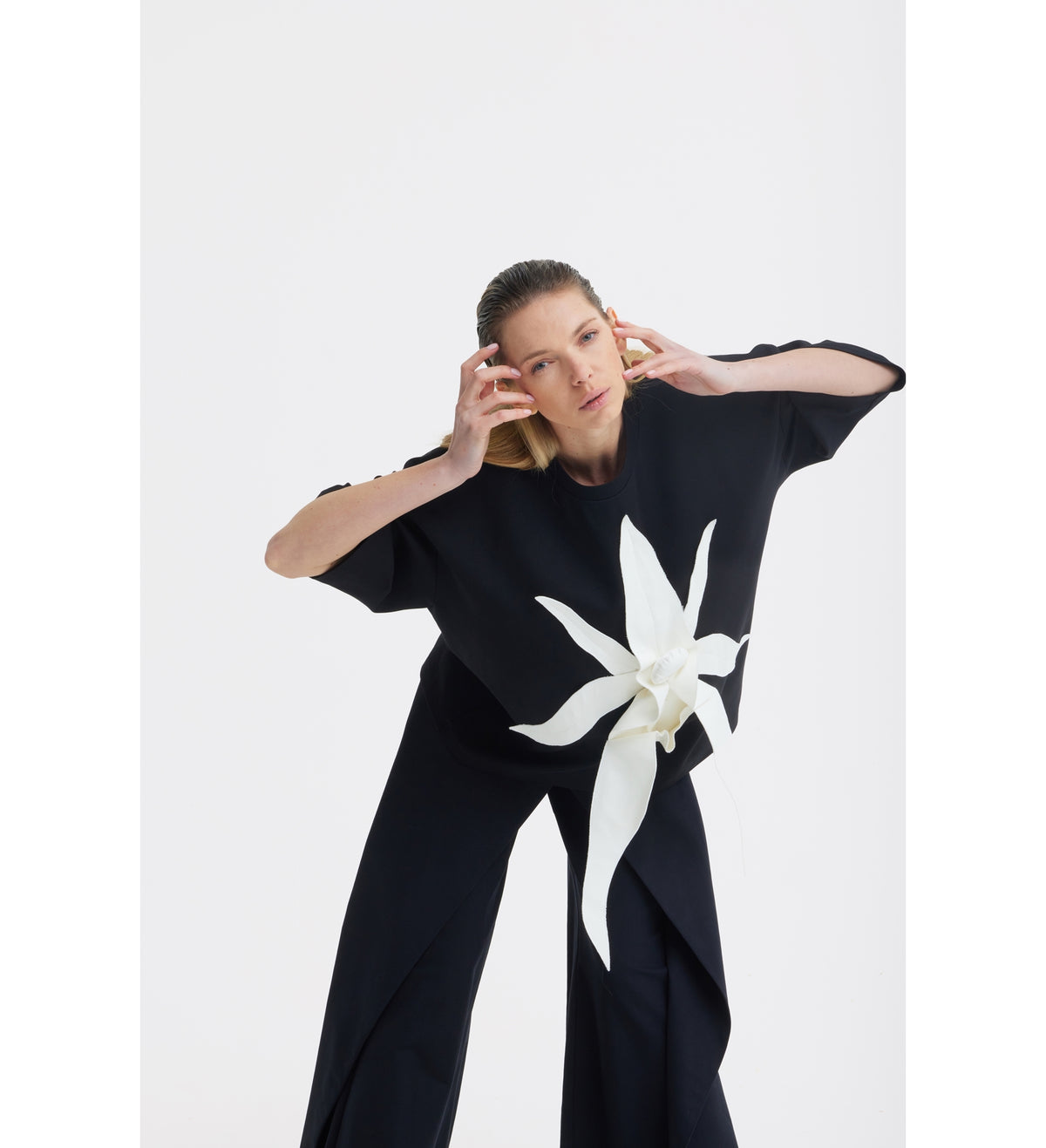 A woman poses against a white background in a JULIA ALLERT Relaxed Fit Knit Top - Black, featuring a sculptural floral appliqué. She leans forward, hands near her head, looking at the camera.
