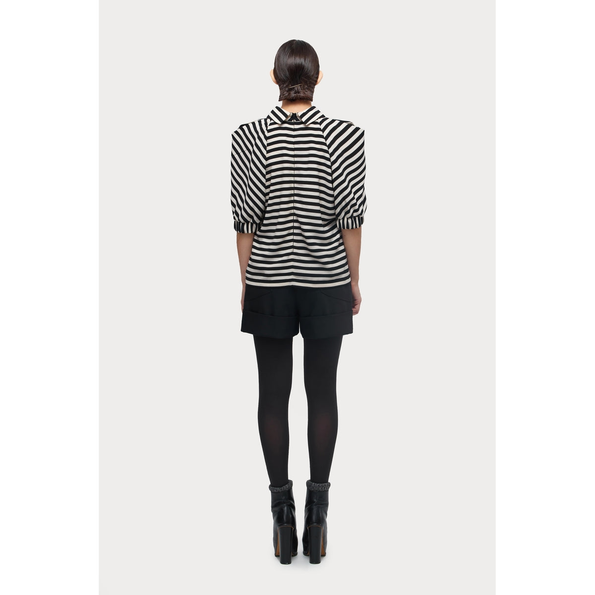 A person stands facing away, wearing the JULIA ALLERT Designer Blouse With Black White Stripes, paired with black shorts, tights, and heeled shoes against a plain light background.