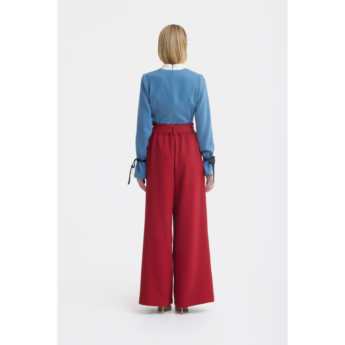 A person stands facing away, wearing a light blue long-sleeve blouse with black accents and JULIA ALLERT Belted High Waist Wide Leg Pants in burgundy. The background is plain white.