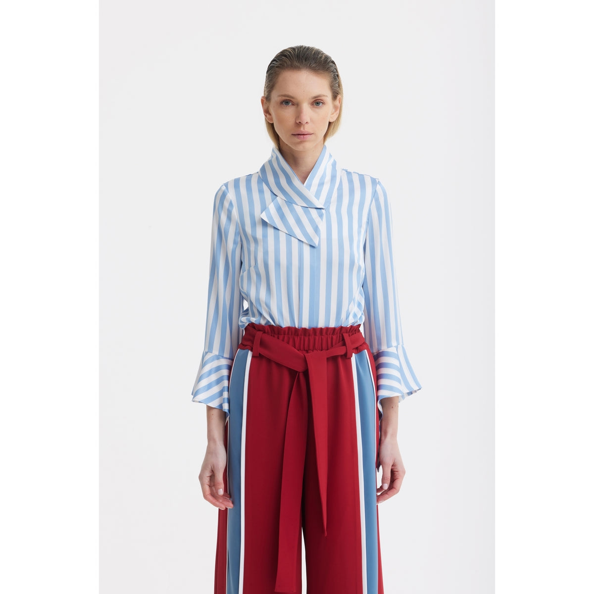 A woman faces forward, wearing a light blue and white striped blouse with flared sleeves, paired with JULIA ALLERT's Belted High Waist Wide Leg Pants in burgundy featuring a paperbag waist and matching blue stripes.