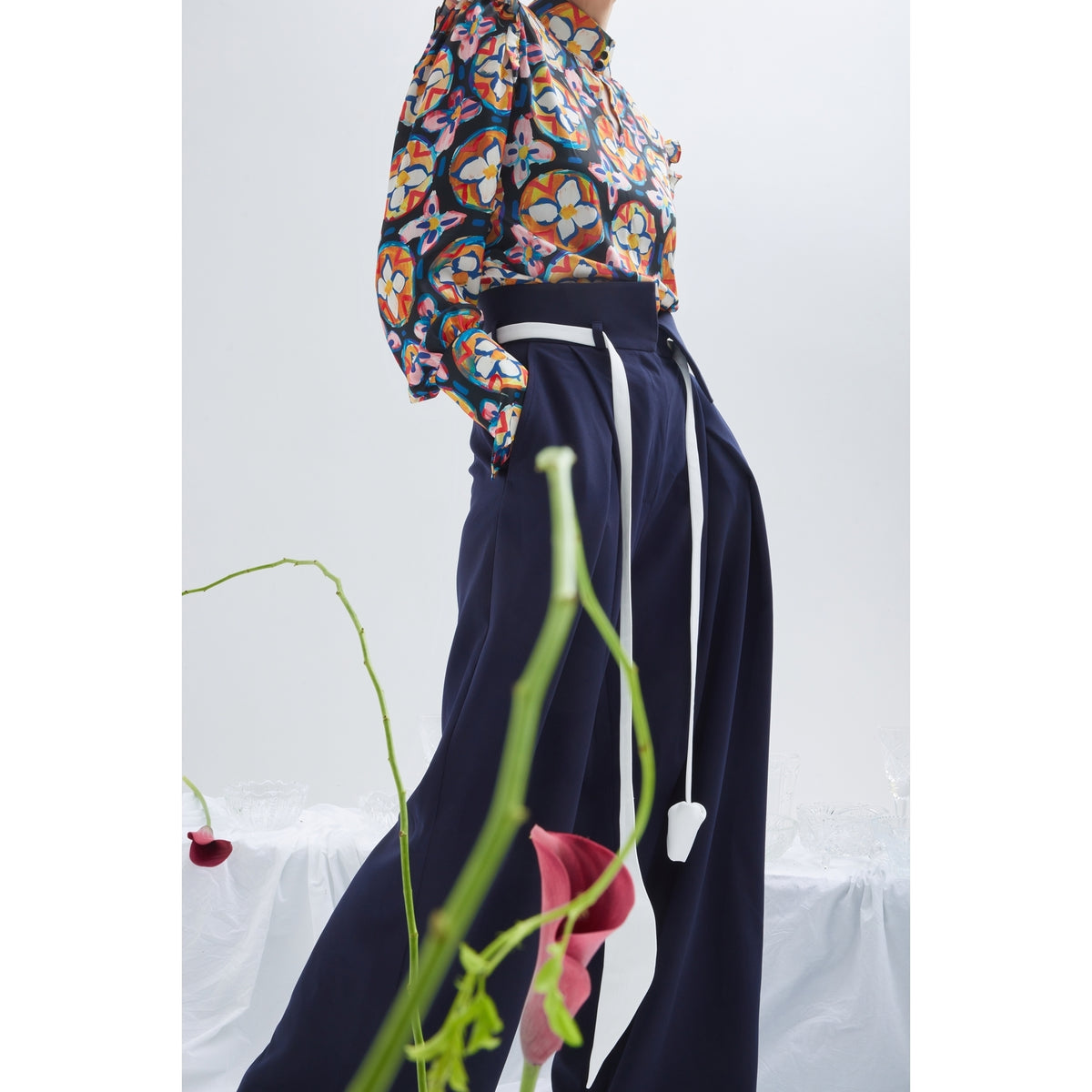 A person wears JULIA ALLERT's Fashion Forward Wide-Leg Pants &amp; Contrast Belt in navy, paired with a vibrant floral blouse. Blurred pink flowers appear in the foreground against a plain, light background.