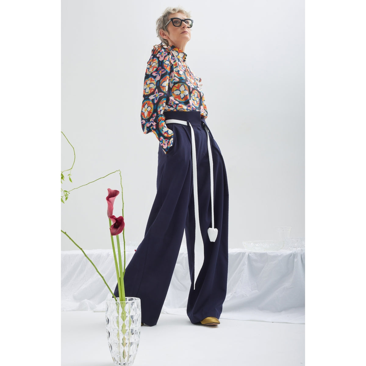 A stylish person in JULIA ALLERT’s Fashion Forward Wide-Leg Pants &amp; Contrast Belt - Navy stands confidently beside a glass vase with two calla lilies against a minimal white background.