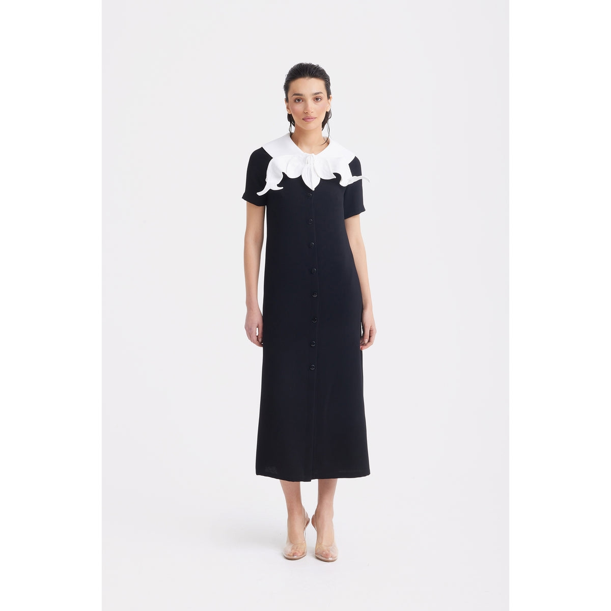 A woman wearing the JULIA ALLERT Straight Midi Shirt Dress With Adjustable Collar in black stands facing forward against a white background. She pairs it with nude heels and has her dark hair pulled back.