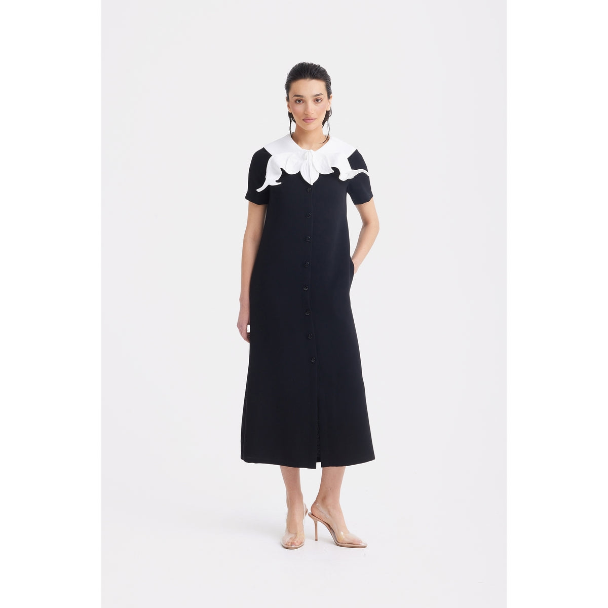 A woman stands against a white background wearing the JULIA ALLERT Straight Midi Shirt Dress With Adjustable Collar - Black, featuring short sleeves and a large adjustable collar. She pairs it with nude heels and keeps her hands in her pockets.