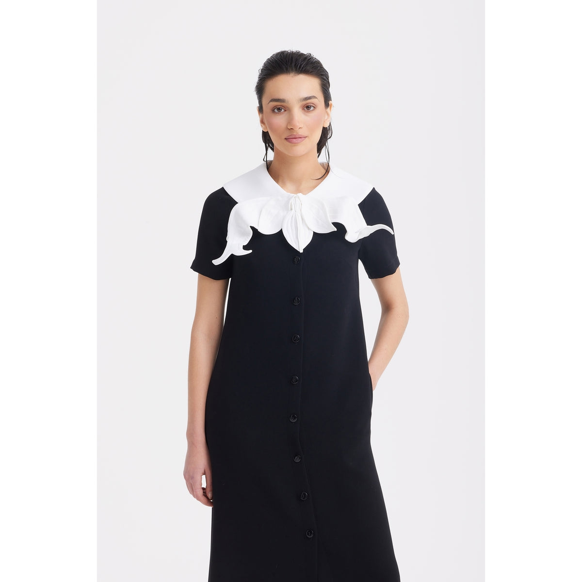 A woman stands against a plain white background wearing the JULIA ALLERT Straight Midi Shirt Dress With Adjustable Collar in black, featuring short sleeves and a large, decorative flower petal-inspired white adjustable collar.