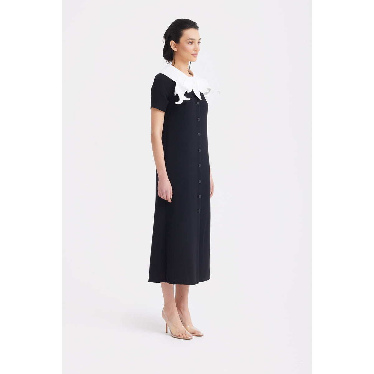 A woman in profile wears the JULIA ALLERT Straight Midi Shirt Dress With Adjustable Collar - Black, styled with large white ruffles and nude heels, set against a plain white background.