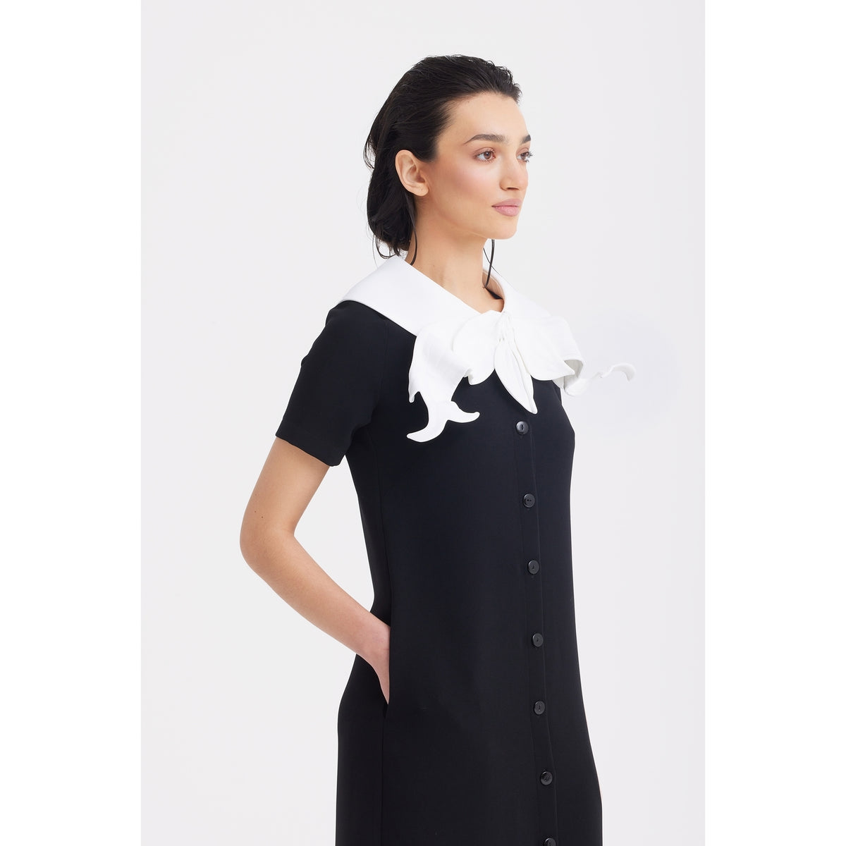 A woman stands sideways, wearing the JULIA ALLERT Straight Midi Shirt Dress With Adjustable Collar in black, featuring a striking sculptural white adjustable collar. Her hands are in her pockets, set against a plain white background.