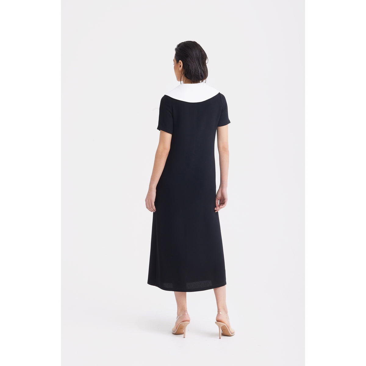 A woman stands with her back to the camera, wearing the JULIA ALLERT Straight Midi Shirt Dress With Adjustable Collar in black and nude heeled sandals, set against a plain white background.