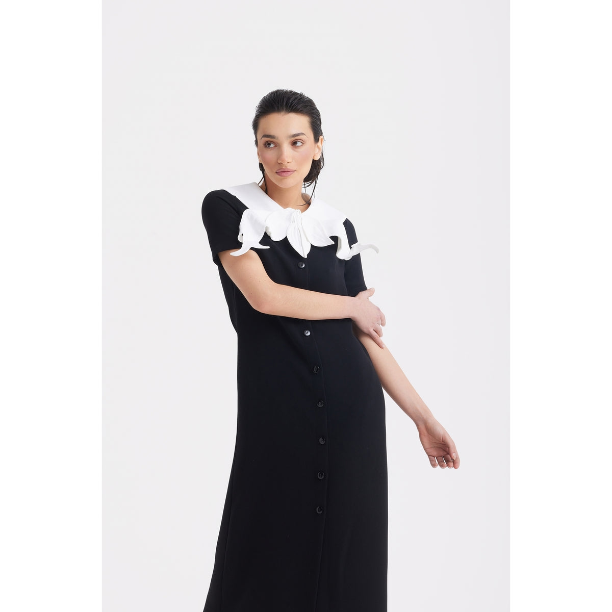 A woman poses against a plain white background in the JULIA ALLERT Straight Midi Shirt Dress With Adjustable Collar—black with short sleeves and a large white ruffled detail. Her dark hair is pulled back.