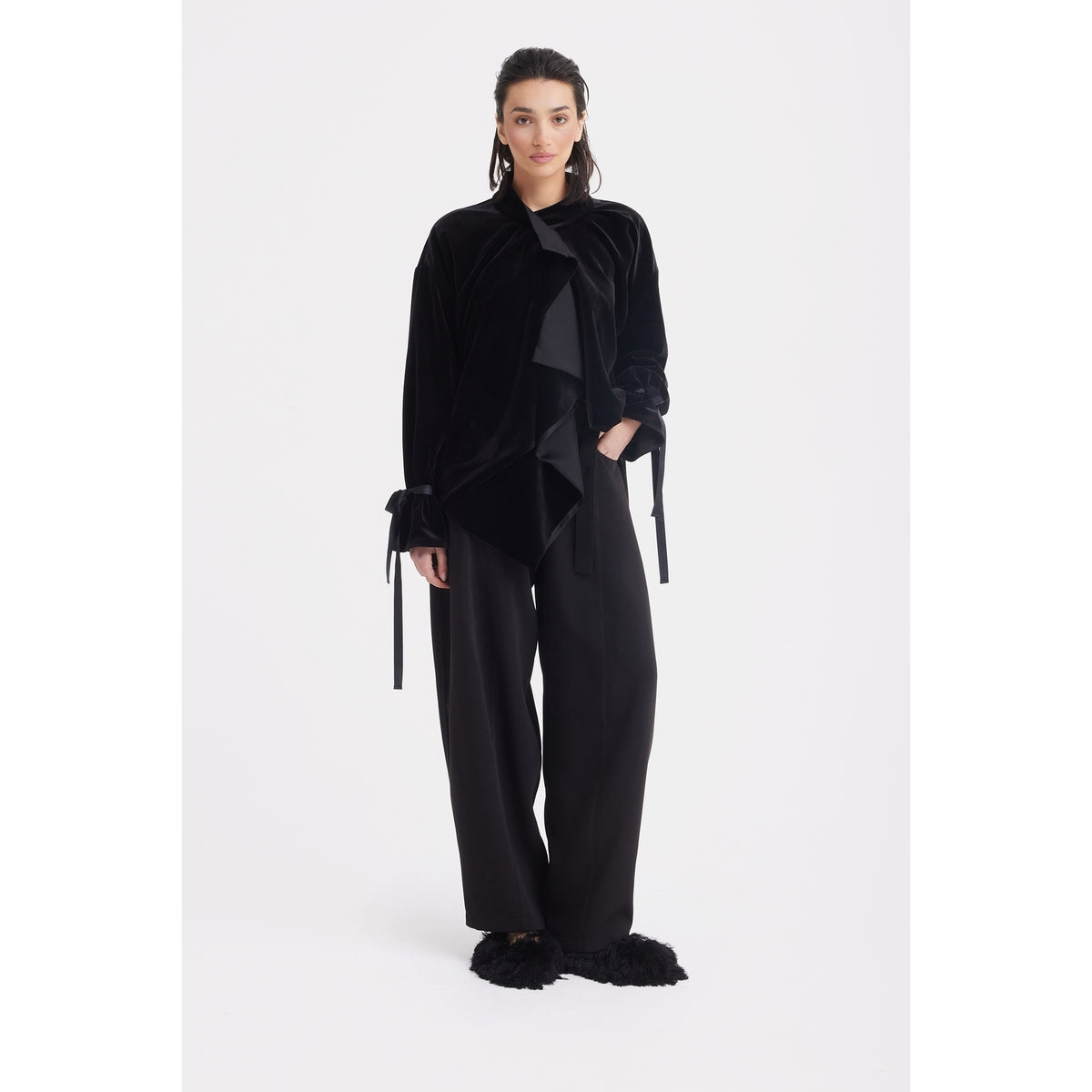 A woman models the JULIA ALLERT Evening Velvet Wrap Jacket With Silk Lining – Black, paired with wide-leg black pants and furry black slippers, standing against a white background.
