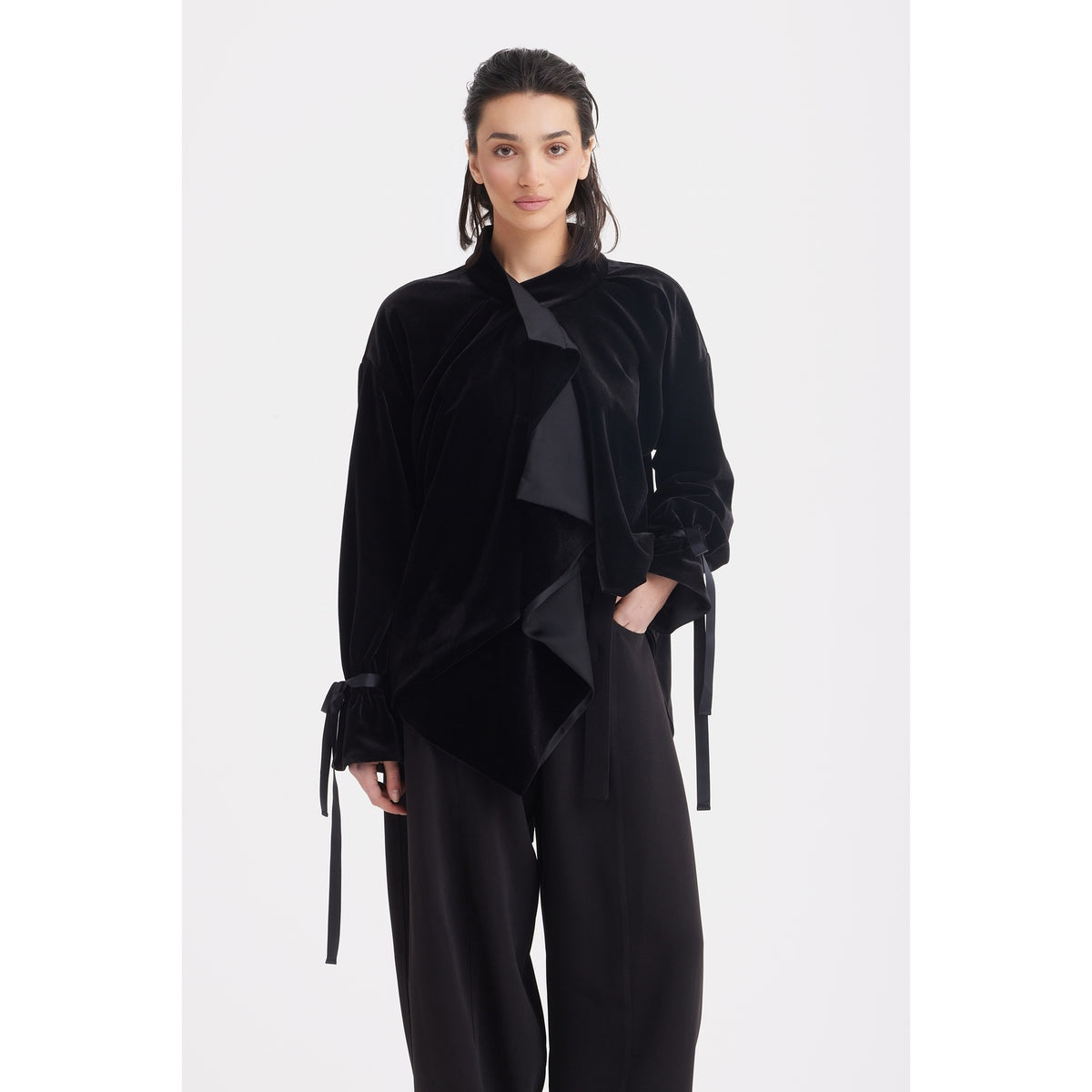 A woman faces forward wearing the JULIA ALLERT Evening Velvet Wrap Jacket With Silk Lining - Black, featuring an asymmetrical ruffled front and wide sleeves tied at the wrists, paired with wide-leg black pants against a plain white background.