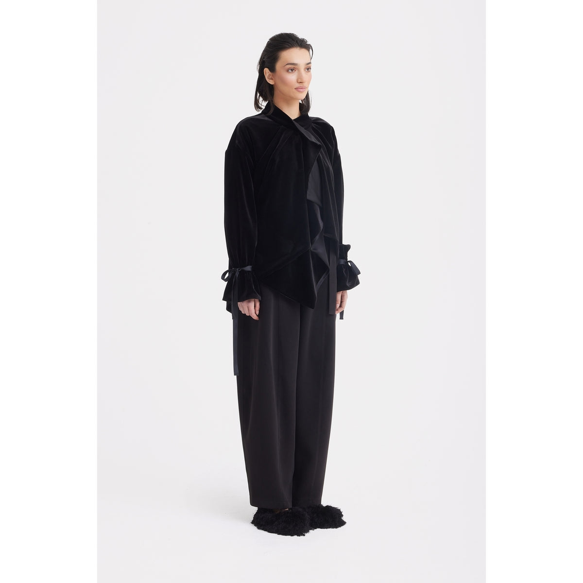 A woman with dark hair pulled back stands against a white background wearing loose black pants and the JULIA ALLERT Evening Velvet Wrap Jacket With Silk Lining - Black, which features oversized sleeves, paired with black furry slippers.