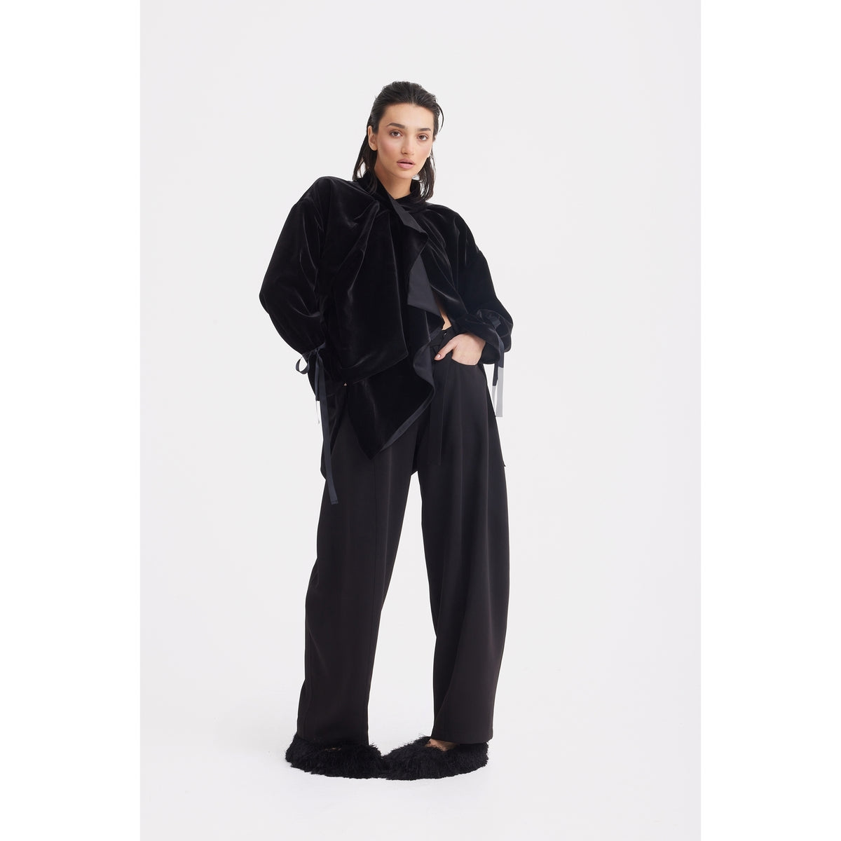 A woman poses confidently against a white background, wearing the JULIA ALLERT Evening Velvet Wrap Jacket With Silk Lining in black, paired with wide-leg black pants and black furry slippers. Her left hand is in her pocket as she looks at the camera.