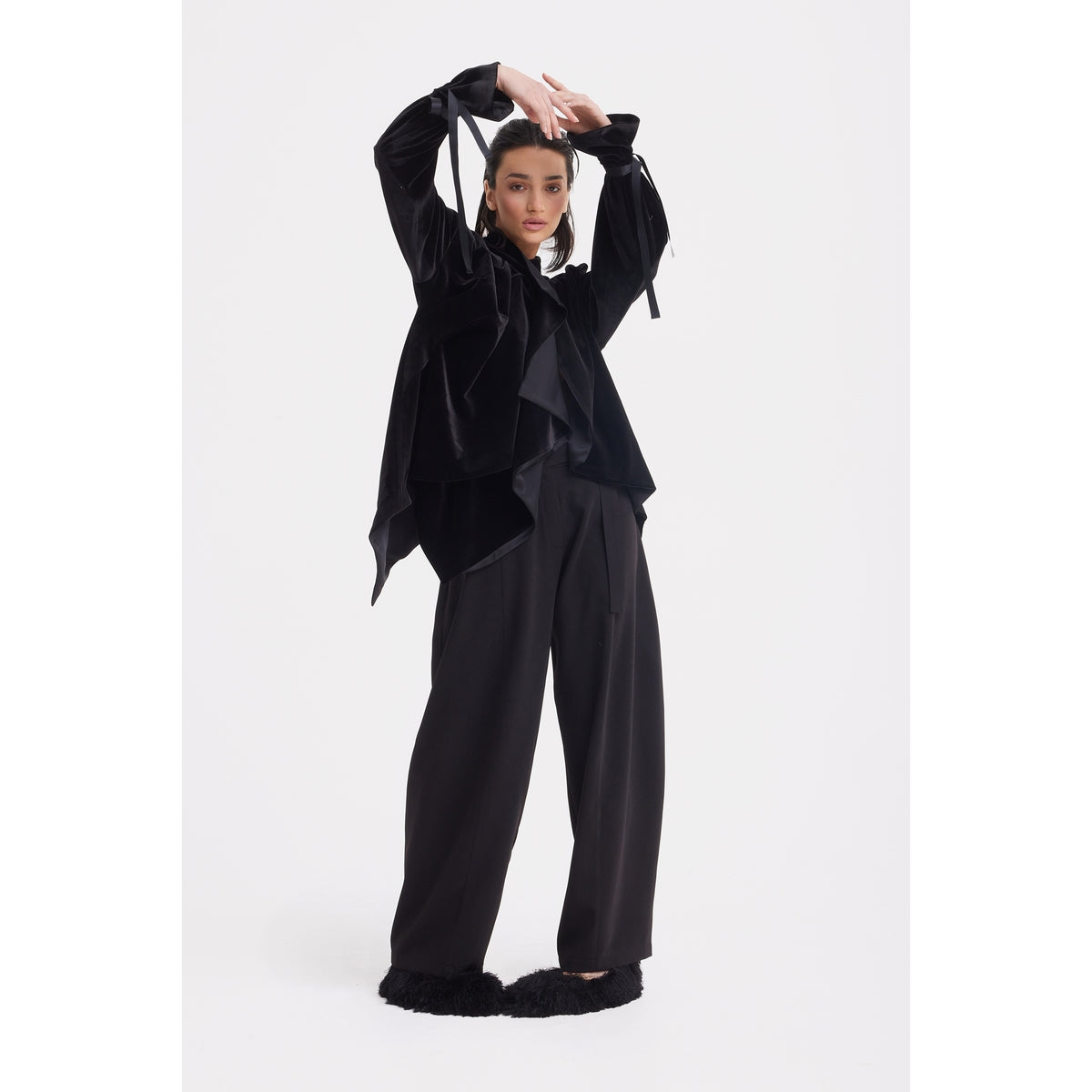 A person poses confidently in a JULIA ALLERT Evening Velvet Wrap Jacket With Silk Lining - Black, paired with wide-leg black trousers and fluffy black shoes, against a plain white background.