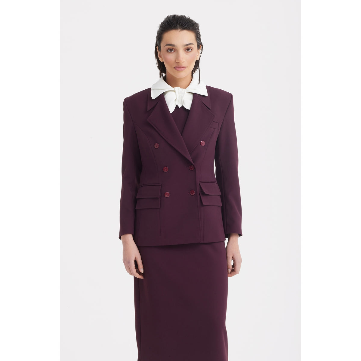 A person with dark hair models the JULIA ALLERT Classic Tailored Double-Breasted Blazer in burgundy, styled as office wear with flap pockets and a large white bow tie, standing against a plain white background.