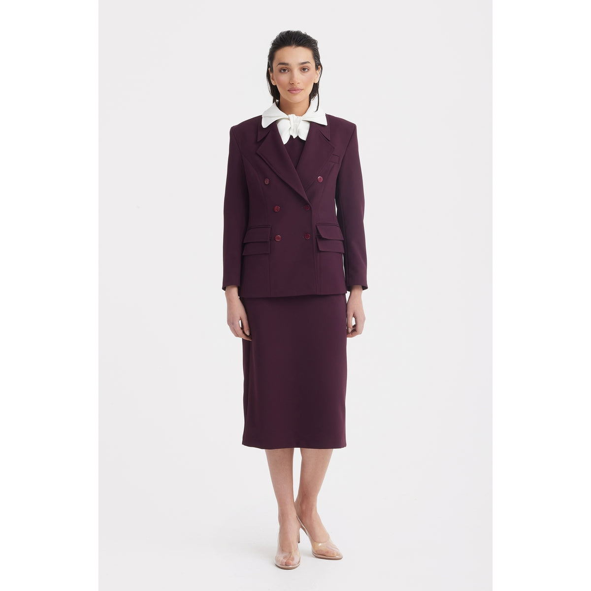 A woman faces forward in the JULIA ALLERT Classic Tailored Double-Breasted Blazer in burgundy, paired with a matching midi skirt, white collared shirt with an oversized bow, and clear heels against a plain white background.