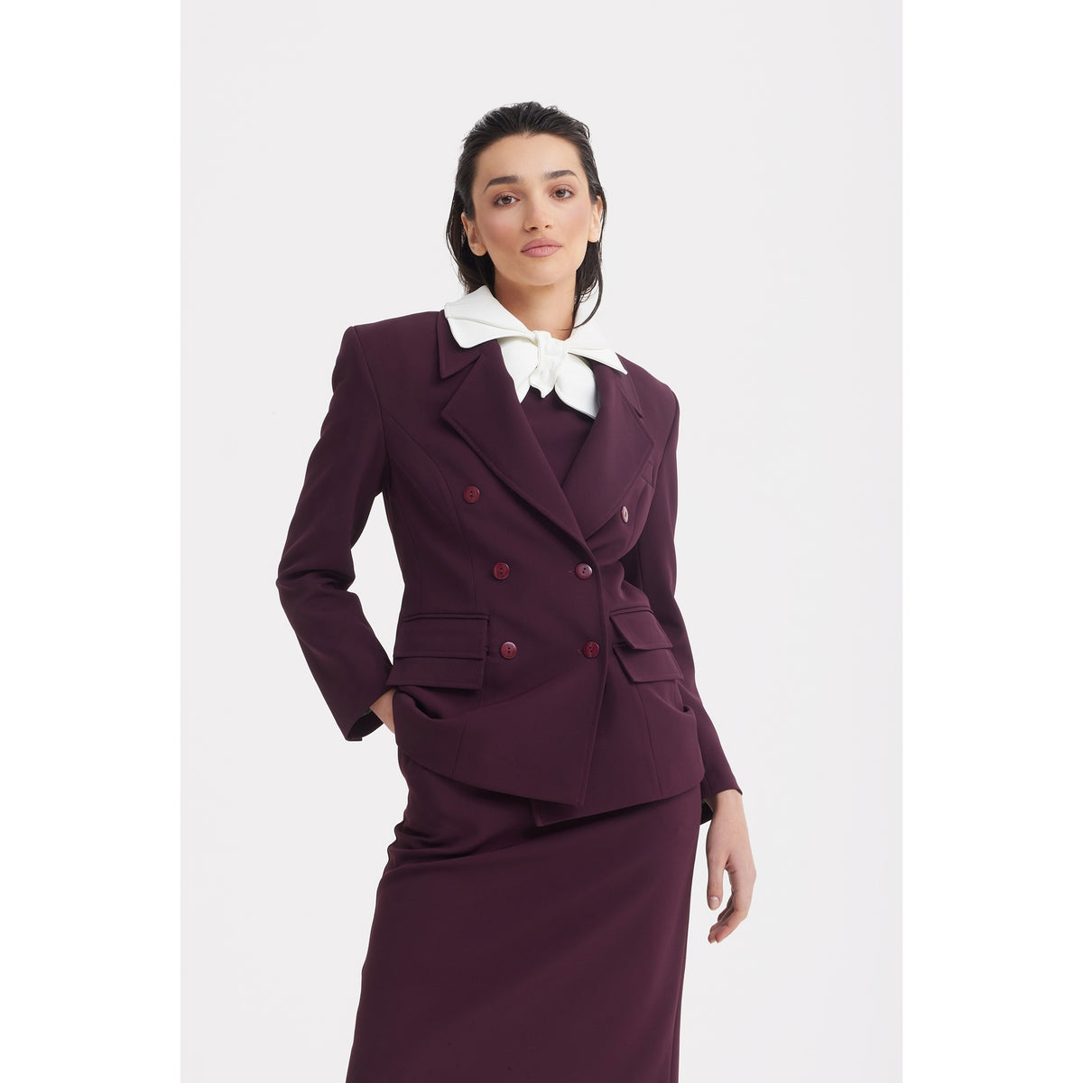 A woman stands confidently against a white background in JULIA ALLERT’s Classic Tailored Double-Breasted Blazer in burgundy, paired with a matching skirt and bold white neck bow, embodying refined modern office style.