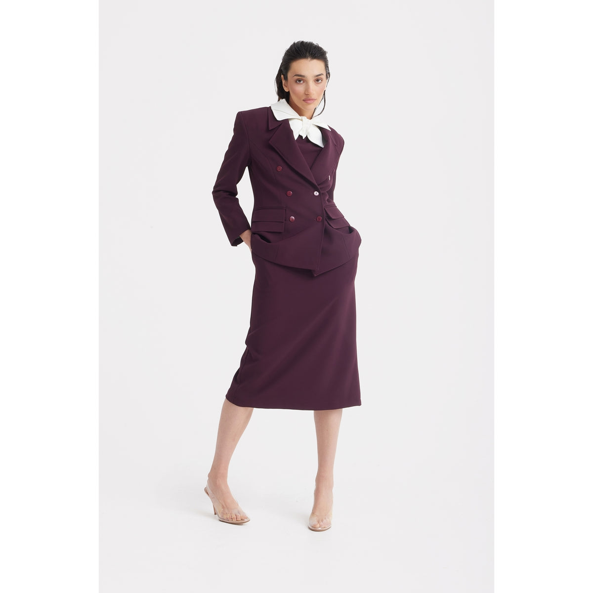 A person stands confidently in the JULIA ALLERT Classic Tailored Double-Breasted Blazer - Burgundy, paired with a matching skirt and white blouse, hands in pockets and legs crossed, highlighting modern office style against a white background.