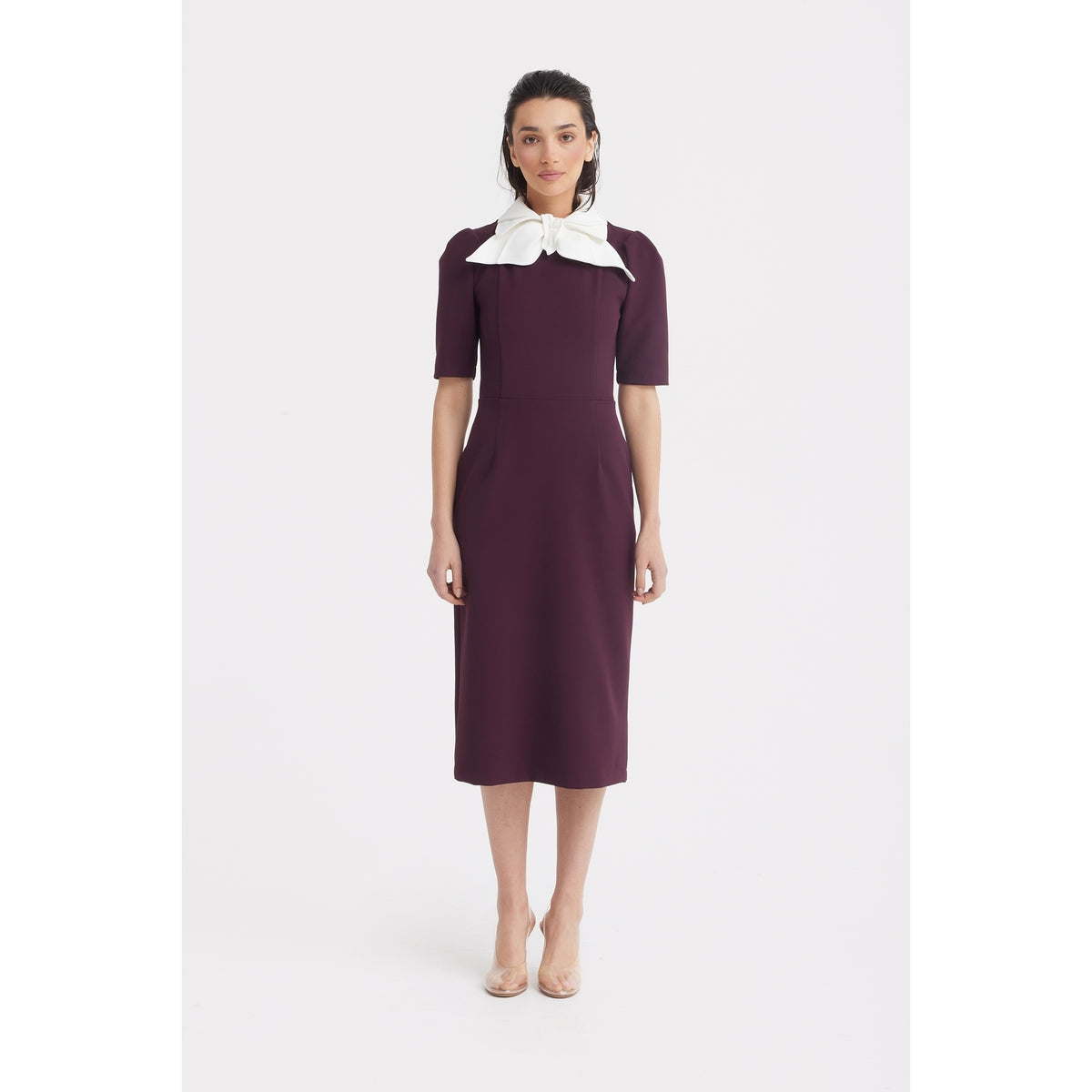 A woman faces forward in the Elegant Midi Dress With Contrast Bow Collar - Burgundy by JULIA ALLERT. The piece features a striking bow collar. She wears neutral makeup, her hair loose, and nude heels against a plain white background.