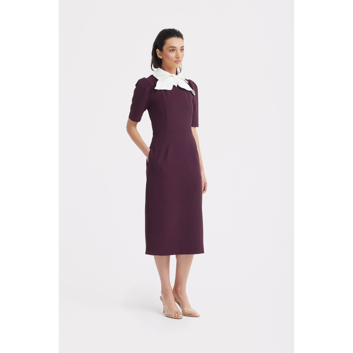 A woman stands in a studio wearing the JULIA ALLERT Elegant Midi Dress With Contrast Bow Collar in burgundy, featuring short sleeves and a contrast bow collar. She pairs it with nude heels, hands in pockets, gazing slightly to the side.