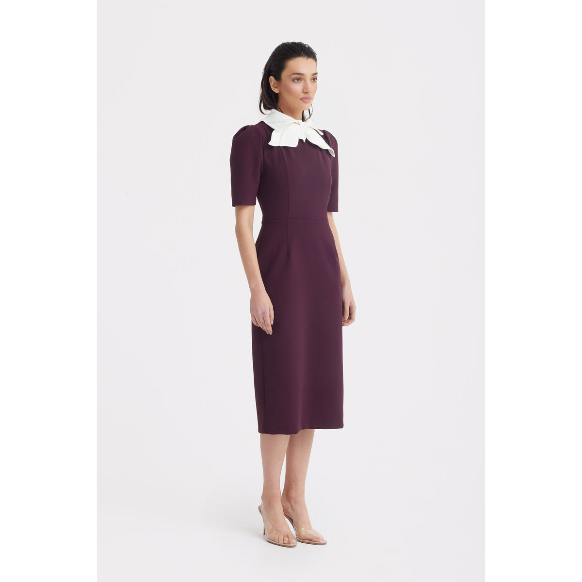 A woman models the JULIA ALLERT Elegant Midi Dress With Contrast Bow Collar in burgundy, paired with clear heels, posing against a plain white background.