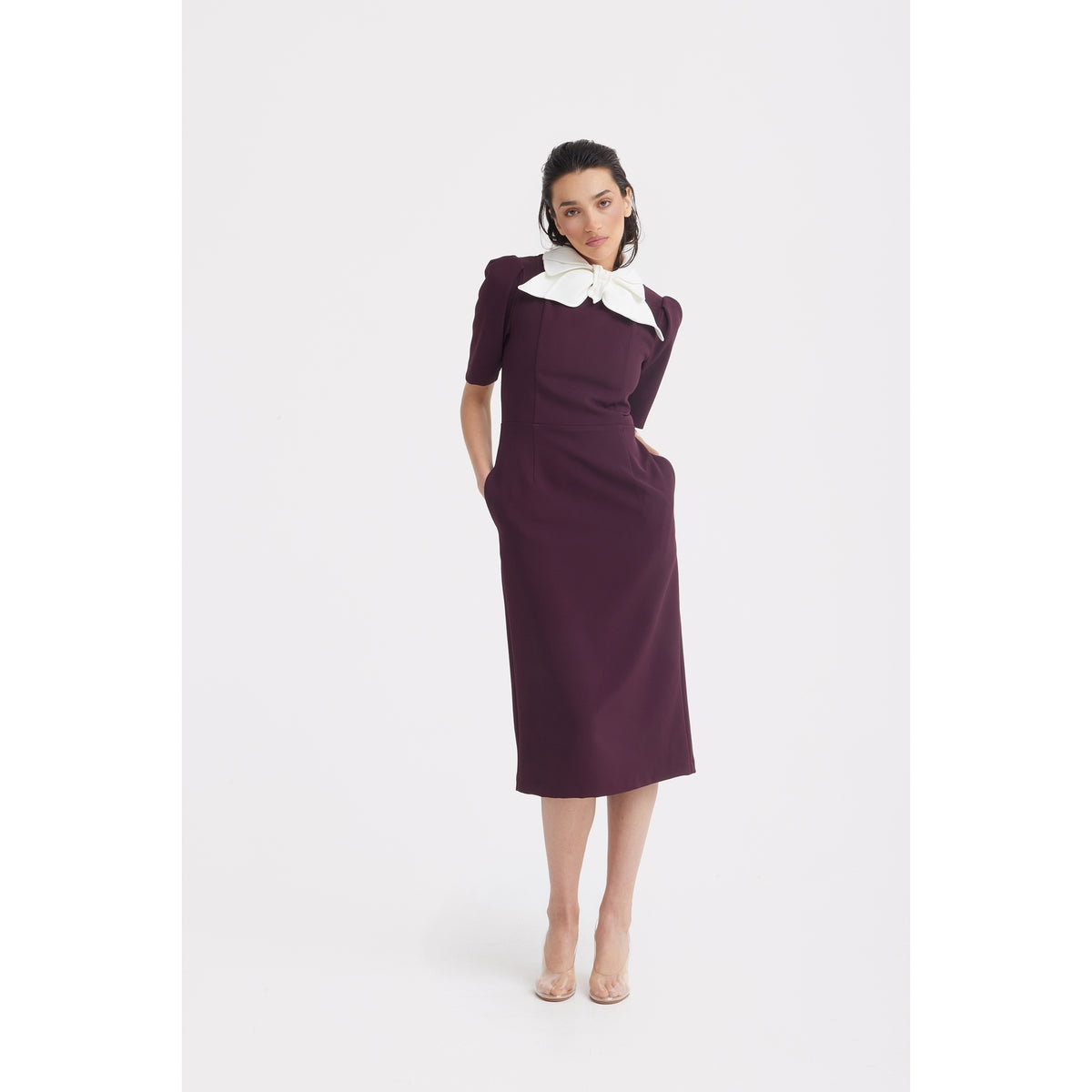 A woman stands against a white background wearing the JULIA ALLERT Elegant Midi Dress With Contrast Bow Collar in burgundy, featuring short sleeves and a striking bow collar. She pairs it with clear high-heeled shoes and has her hands in her pockets.