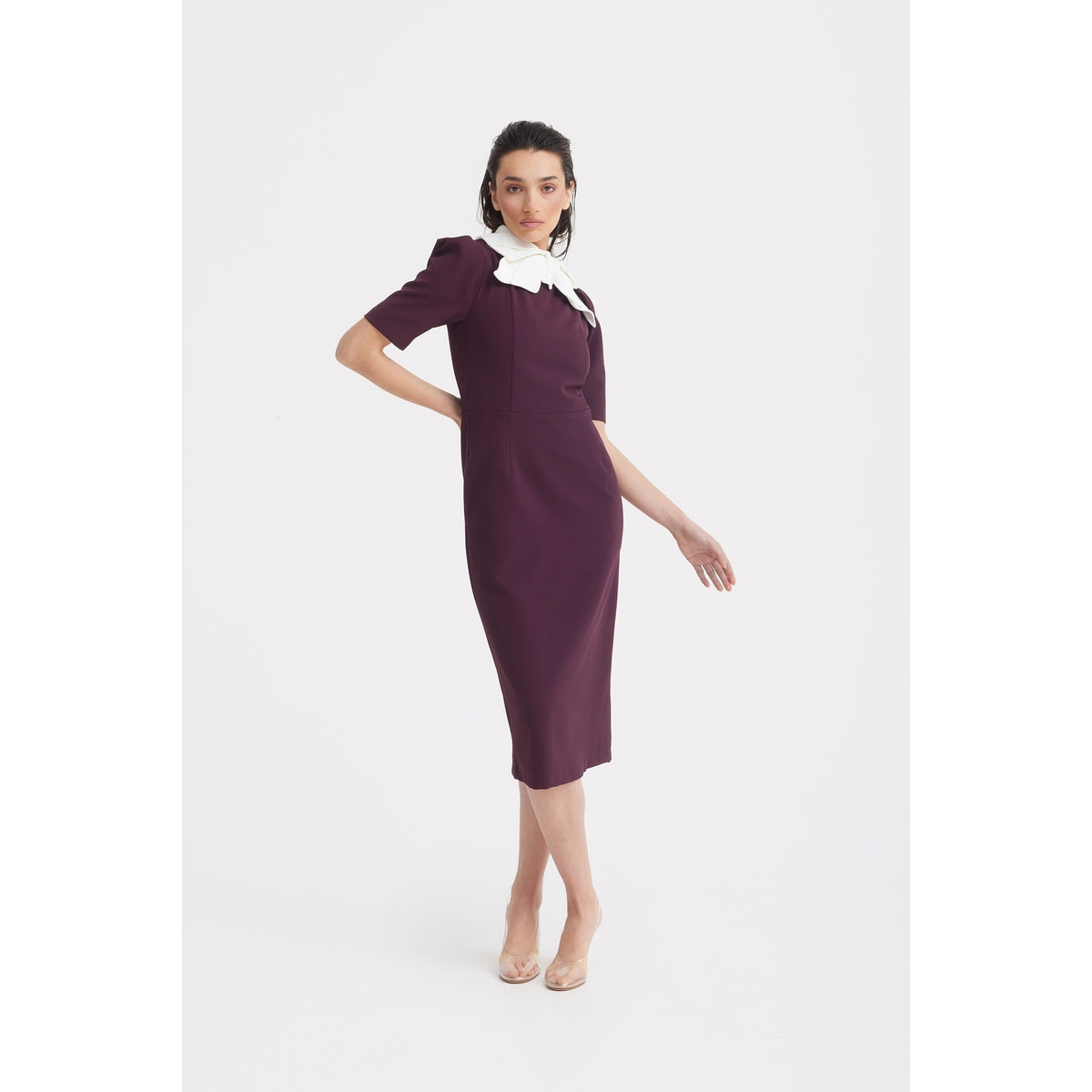 A woman poses in the JULIA ALLERT Elegant Midi Dress With Contrast Bow Collar - Burgundy, standing against a plain white background with clear high heels and her hair styled back.