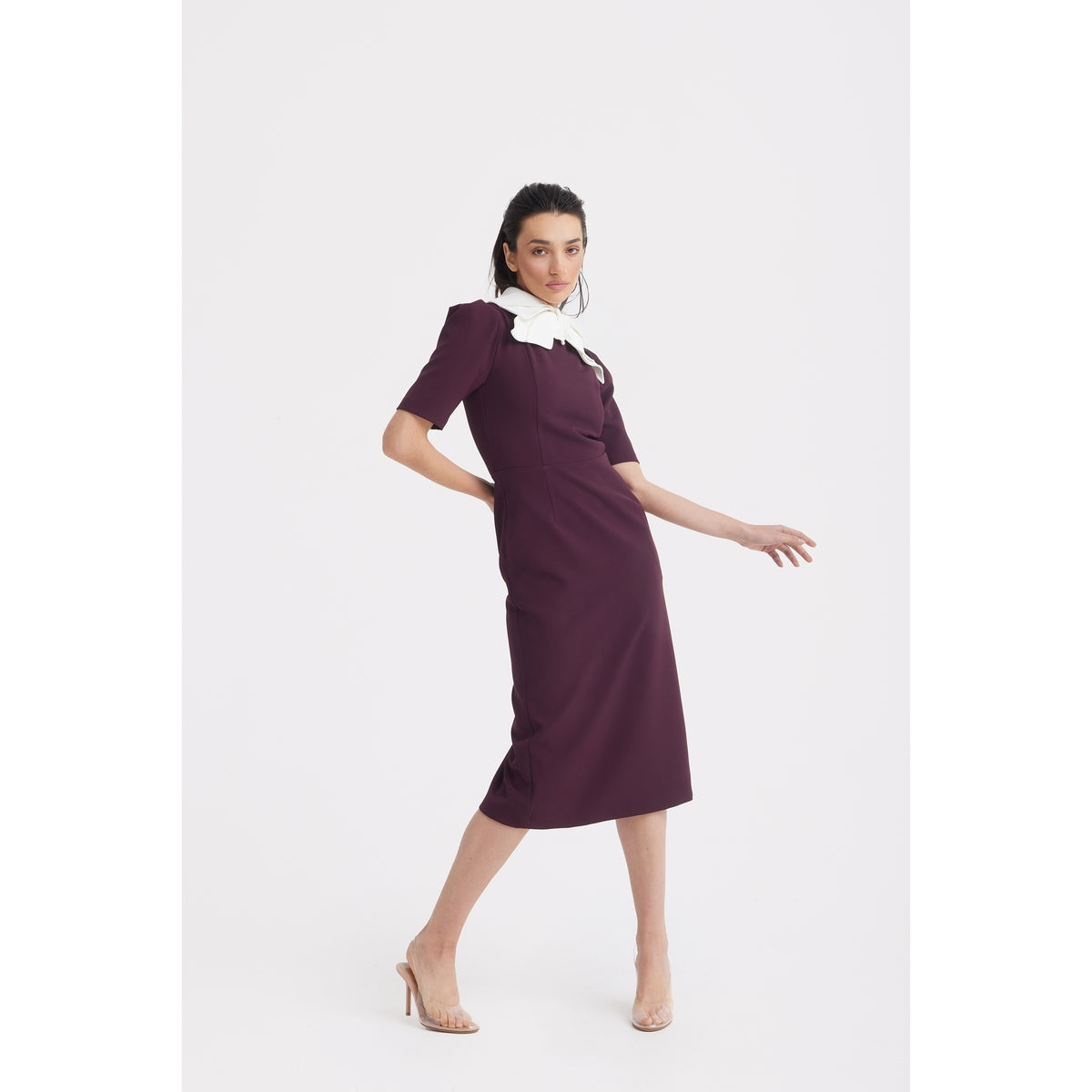 A woman models the JULIA ALLERT Elegant Midi Dress With Contrast Bow Collar in burgundy, a knee-length, short-sleeve style with a standout bow collar, paired with clear heels against a plain white background.