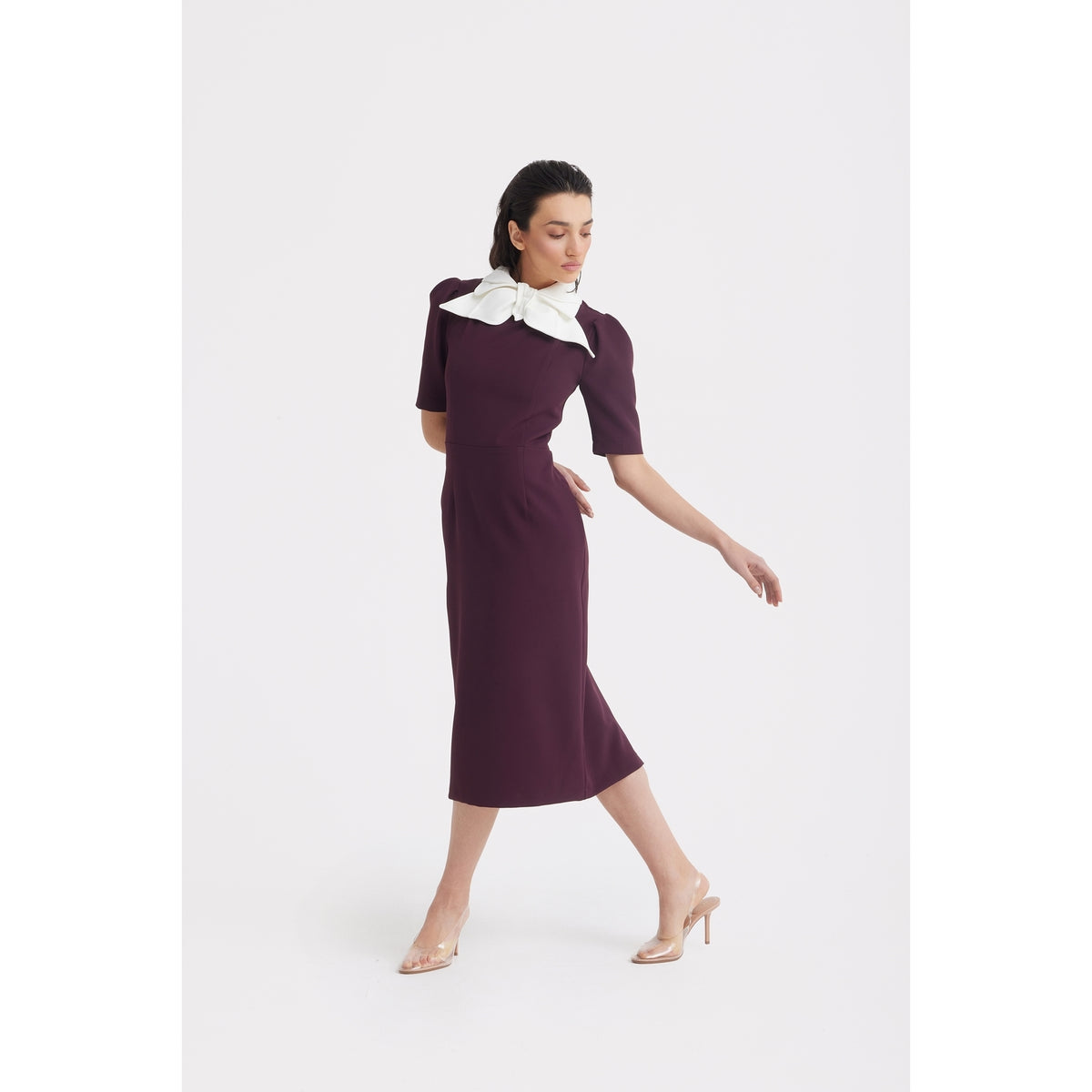 A woman poses in a JULIA ALLERT Elegant Midi Dress With Contrast Bow Collar - Burgundy, featuring short sleeves and a contrasting bow collar, styled with clear high heels against a plain white background.