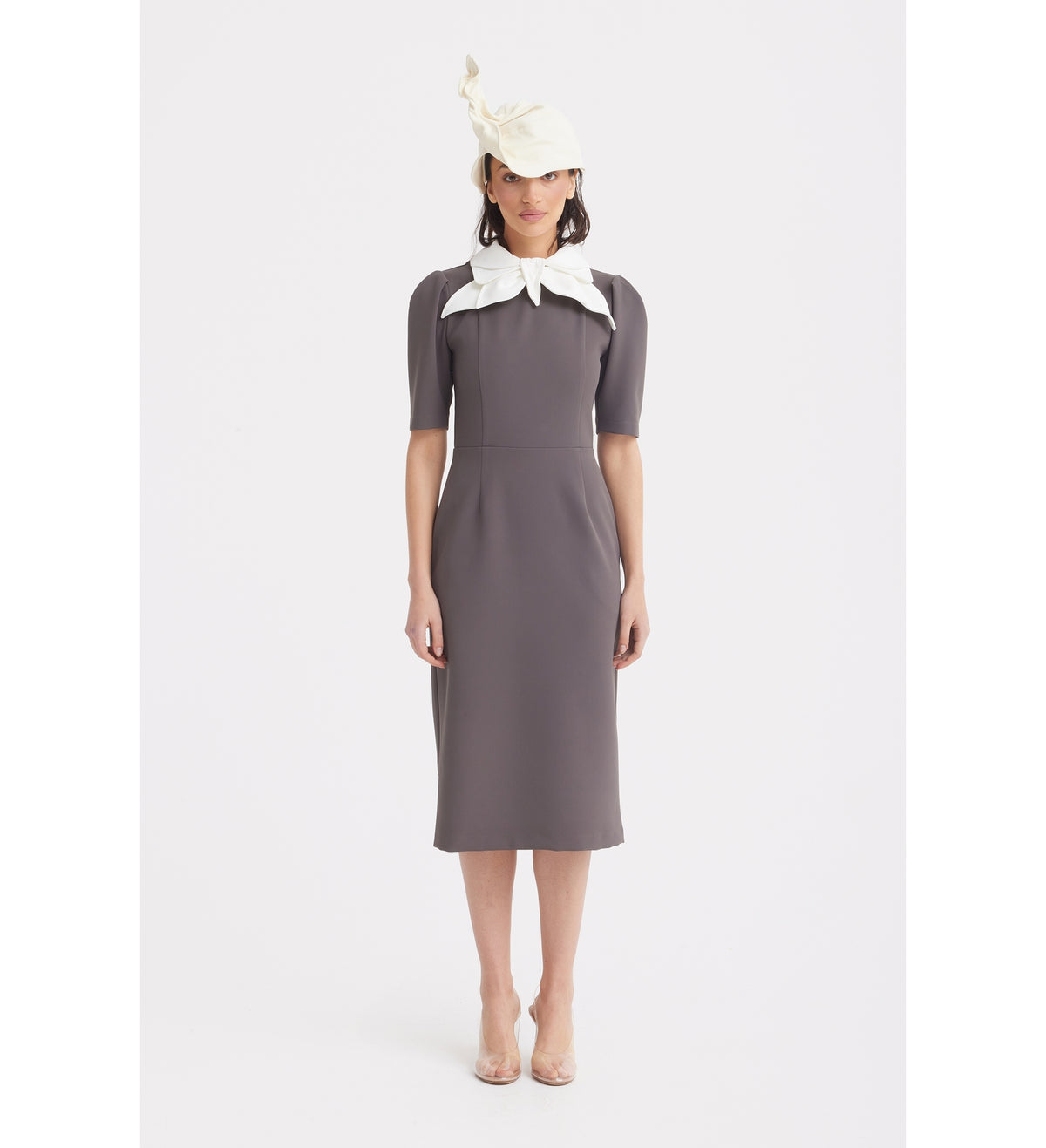 A woman faces forward in the JULIA ALLERT Elegant Midi Dress With Contrast Bow Collar – Taupe, styled with a cream hat and clear heels, standing against a plain white background.