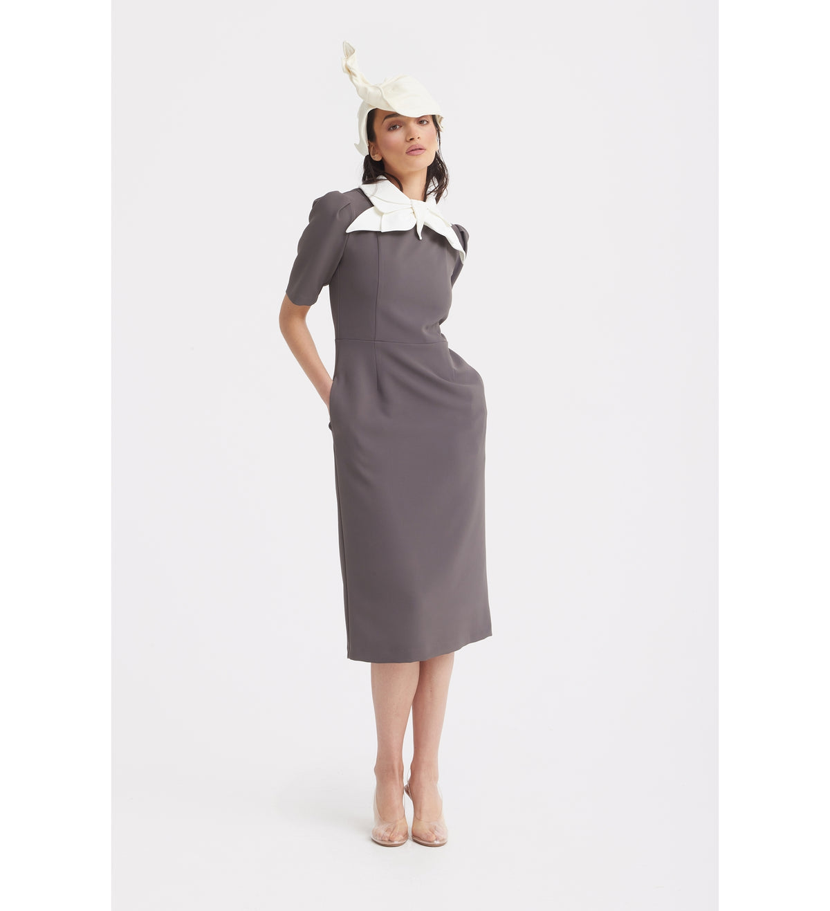 A woman poses in the Elegant Midi Dress With Contrast Bow Collar - Taupe by JULIA ALLERT, featuring a large contrast bow collar, abstract white hat, and clear heels. She stands against a plain white background with her hands in her pockets.