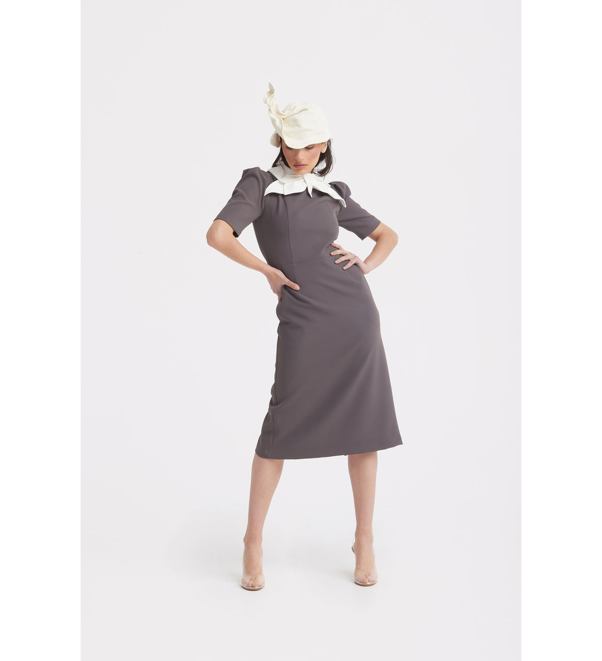 A woman poses confidently in a JULIA ALLERT Elegant Midi Dress With Contrast Bow Collar - Taupe, styled with a cream hat and clear heels, against a plain white background.
