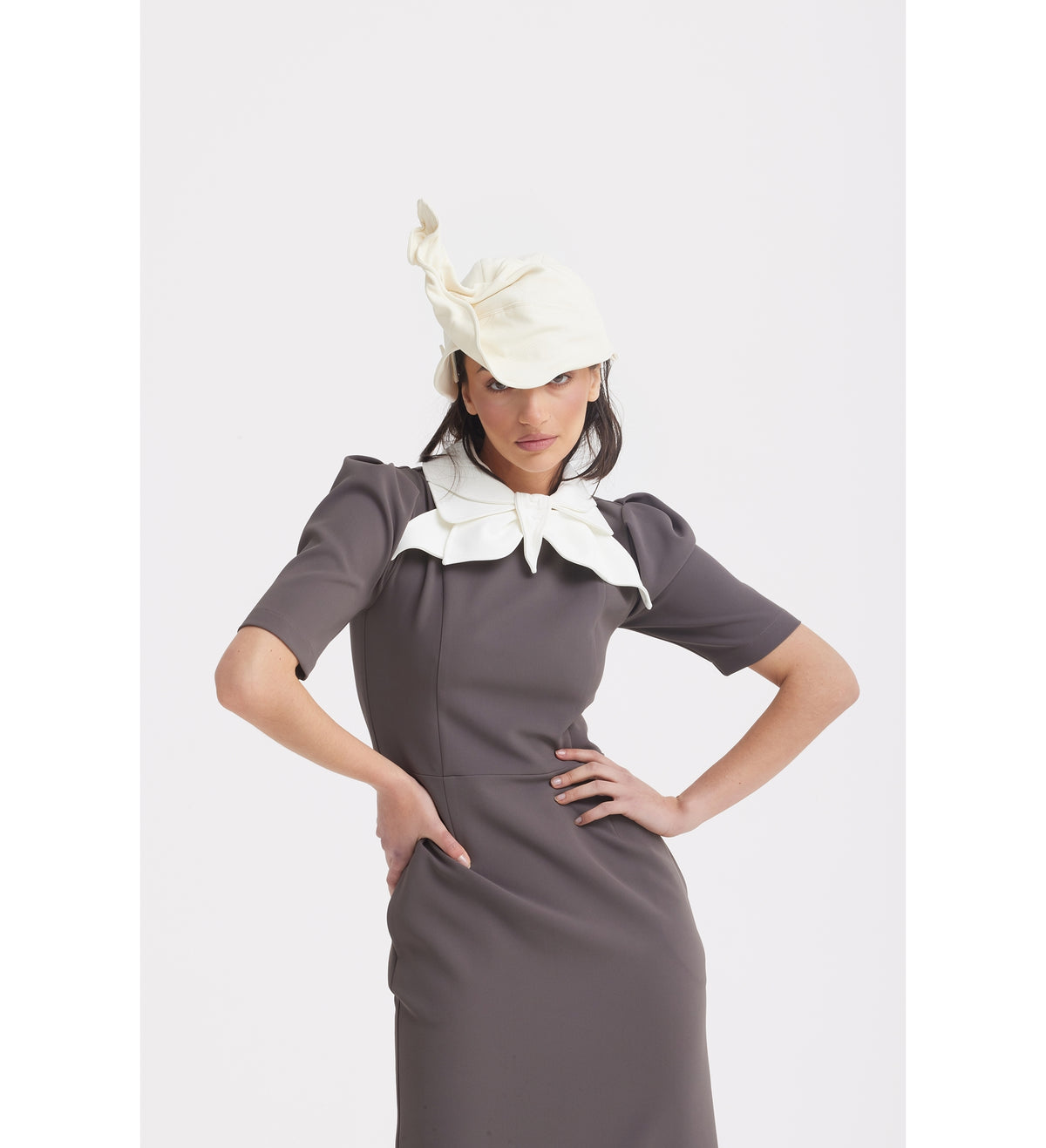 A woman models the JULIA ALLERT Elegant Midi Dress With Contrast Bow Collar in taupe, paired with a chic white hat. She stands against a plain white background with one hand on her hip.