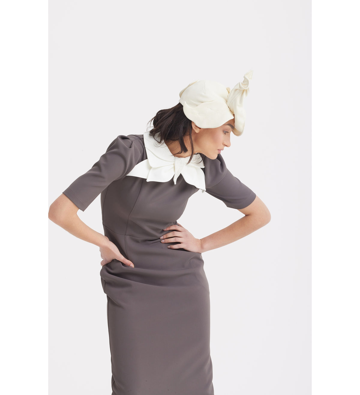 A woman in a fitted taupe Elegant Midi Dress With Contrast Bow Collar by JULIA ALLERT leans forward, wearing a sculptural off-white headpiece against a plain white background.