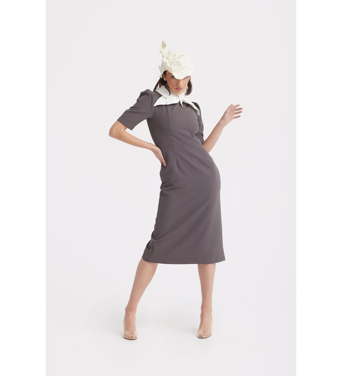 A woman models the JULIA ALLERT Elegant Midi Dress With Contrast Bow Collar - Taupe, featuring short sleeves and a contrast bow collar, paired with a cream hat and clear heels against a plain white background.
