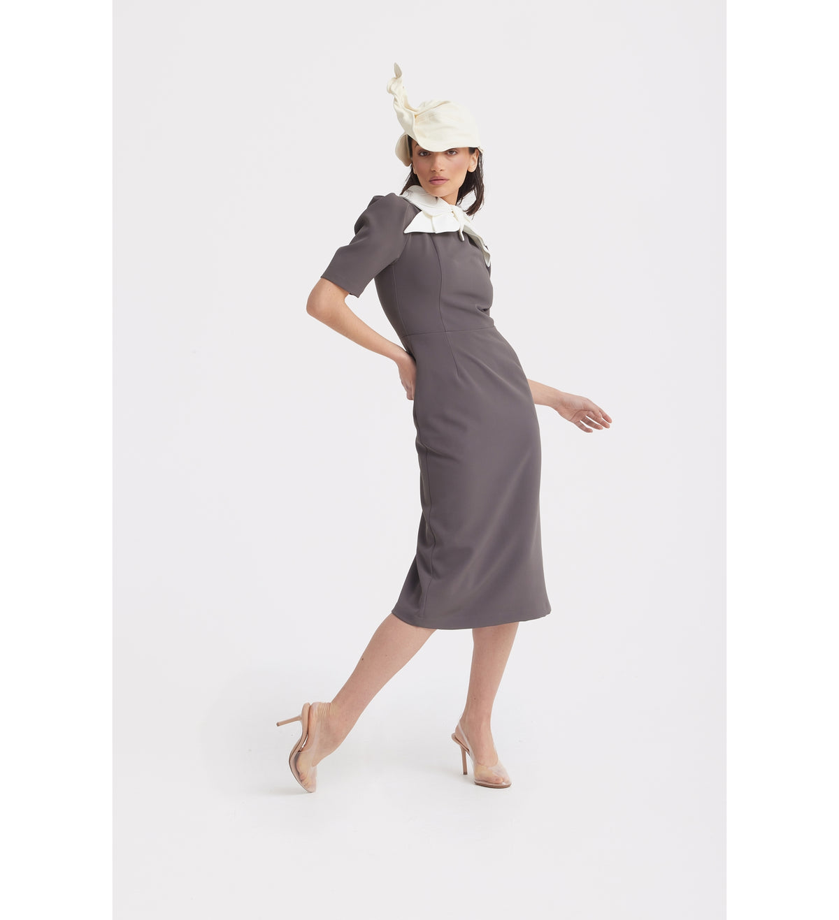A woman poses in the JULIA ALLERT Elegant Midi Dress With Contrast Bow Collar - Taupe, styled with nude heels and a white hat with decorative details, against a plain white background.