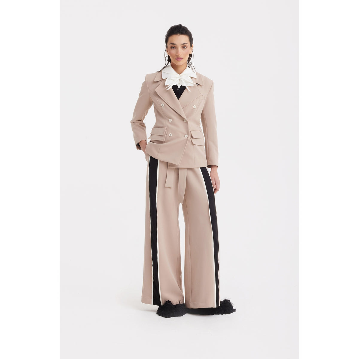 A woman faces forward in a JULIA ALLERT Classic Tailored Double-Breasted Blazer in beige, paired with wide-leg pants featuring black side stripes and feathery hems. She adds a large white ruffled collar and black shoes to complete the look.