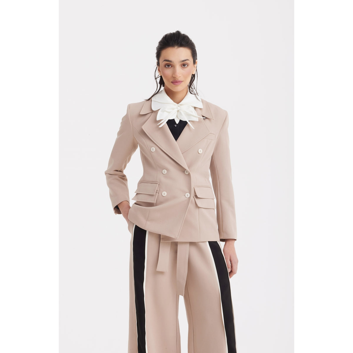 A woman with dark, pulled-back hair and a neutral expression wears the JULIA ALLERT Classic Tailored Double-Breasted Blazer in beige, paired with wide beige pants featuring black side stripes and a large white bow blouse.