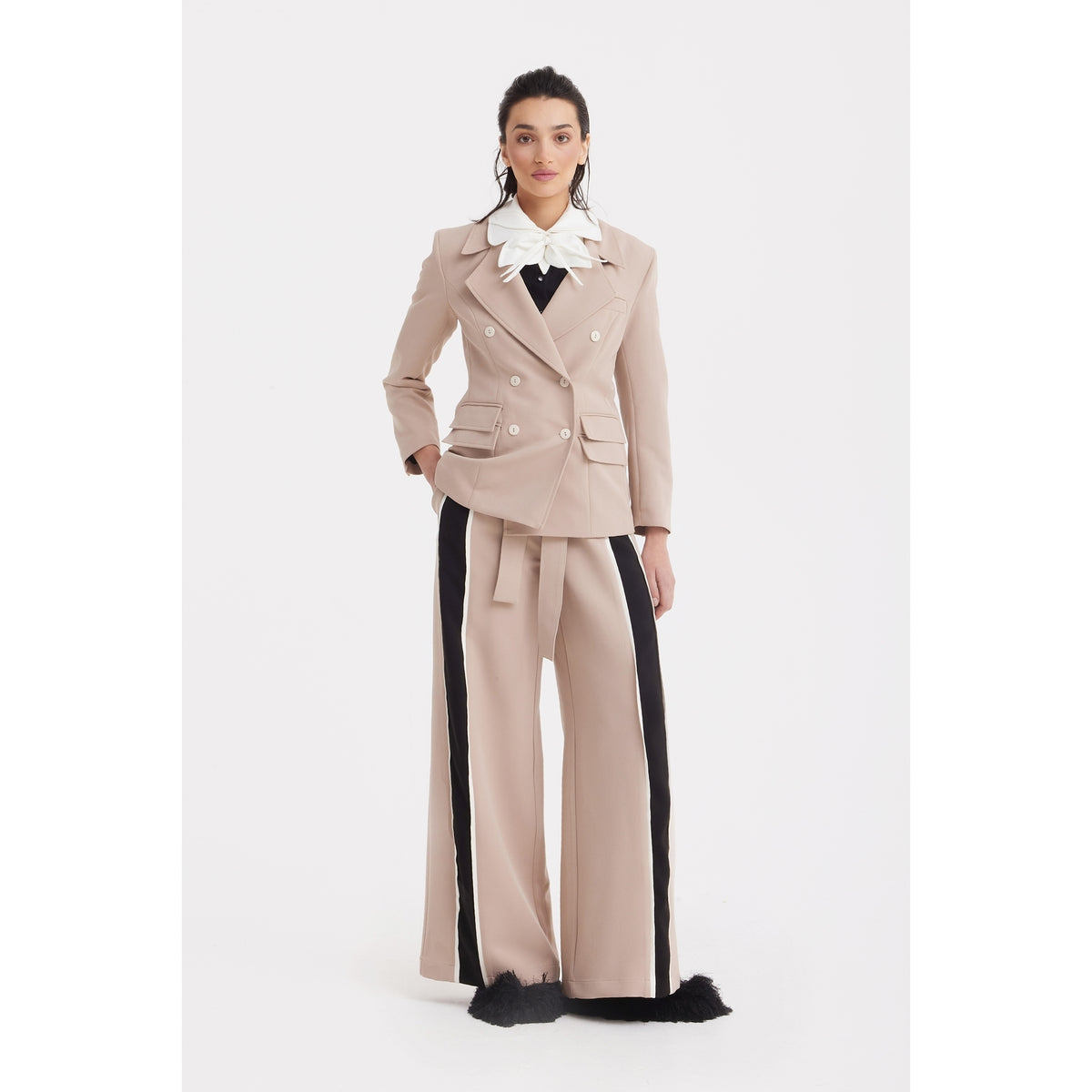 A woman faces forward in a JULIA ALLERT Classic Tailored Double-Breasted Blazer - Beige with matching wide-leg pants featuring black side stripes and furry cuffs, a large white ruffled collar, and pulled-back dark hair against a plain white background.