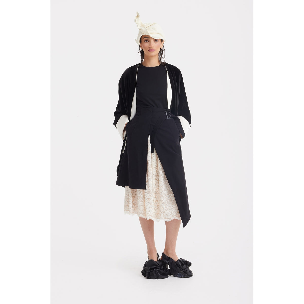 A person stands against a white background wearing the JULIA ALLERT Loose Velvet Cape Jacket With Zip Front in black, styled with a black top, cream lace skirt, ruffled slip-on shoes, and a cream sculptural hat. Hands are in the jacket's pockets.