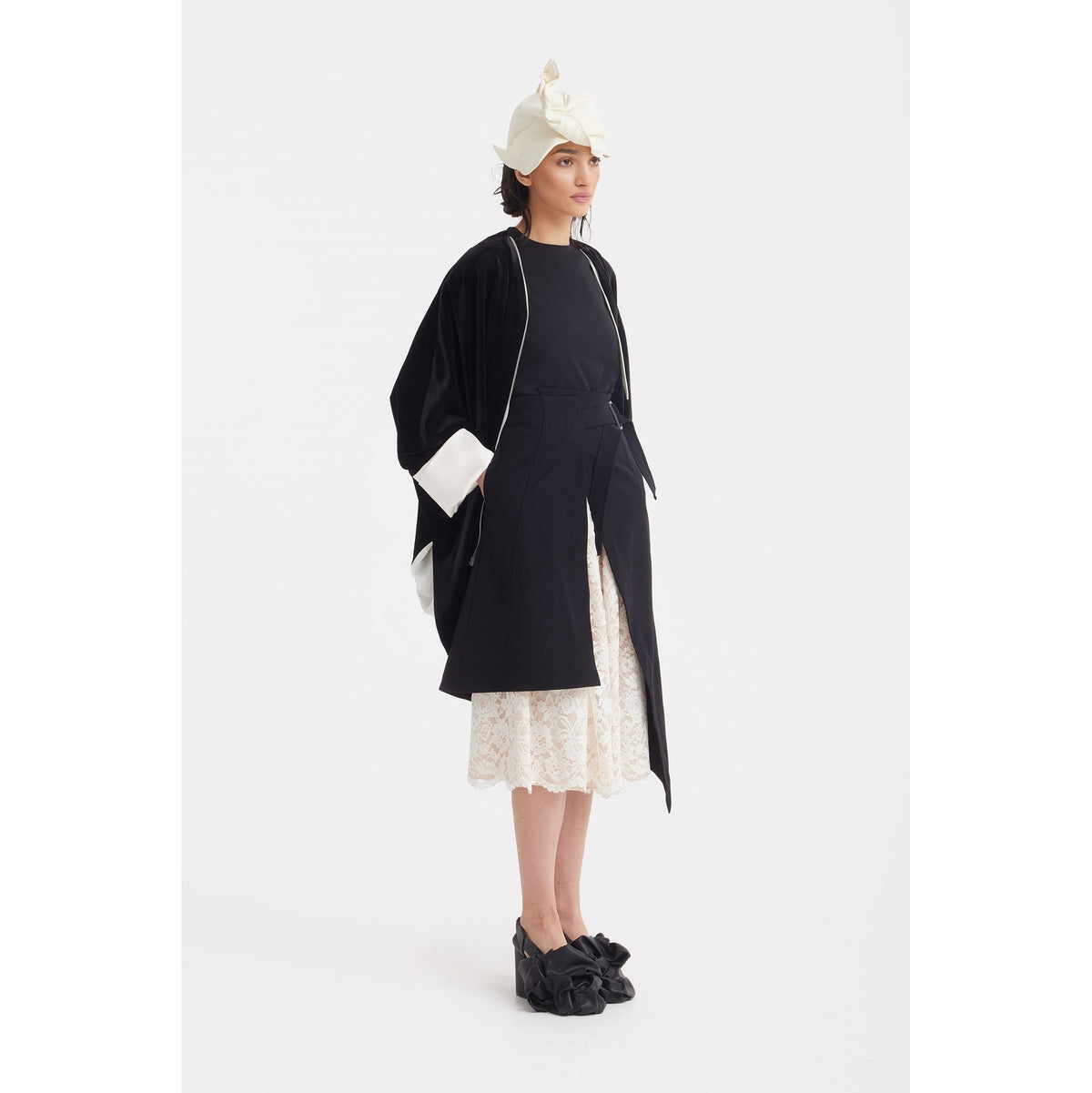 A woman stands against a white background wearing the JULIA ALLERT Loose Velvet Cape Jacket With Zip Front in black, styled with white cuffs, a white lace skirt, black top, black bow shoes, and a knotted white headpiece.